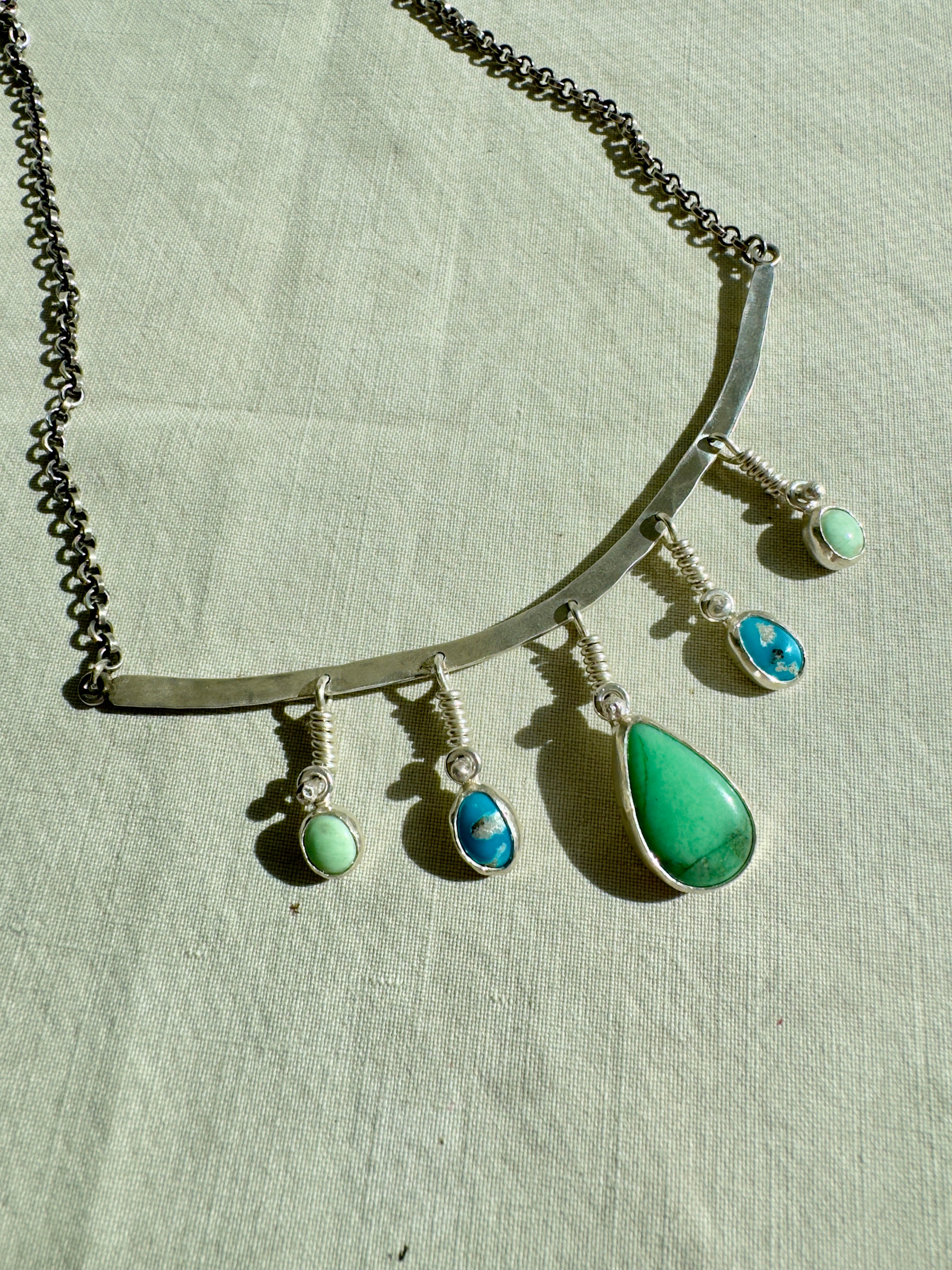 Sterling Silver hammered bar necklace with beautiful green Lucid center stone, flanked by two bright blue castle dome stones and two pale green lucin variscite stones for a total of five stones. The sterling silver bar is attached to a 2.7mm flat rolo chain, oxidized to give it a darker color, with a sterling silver toggle clasp. The necklace rests right on your collarbone, making it the perfect necklace to dress up or down.